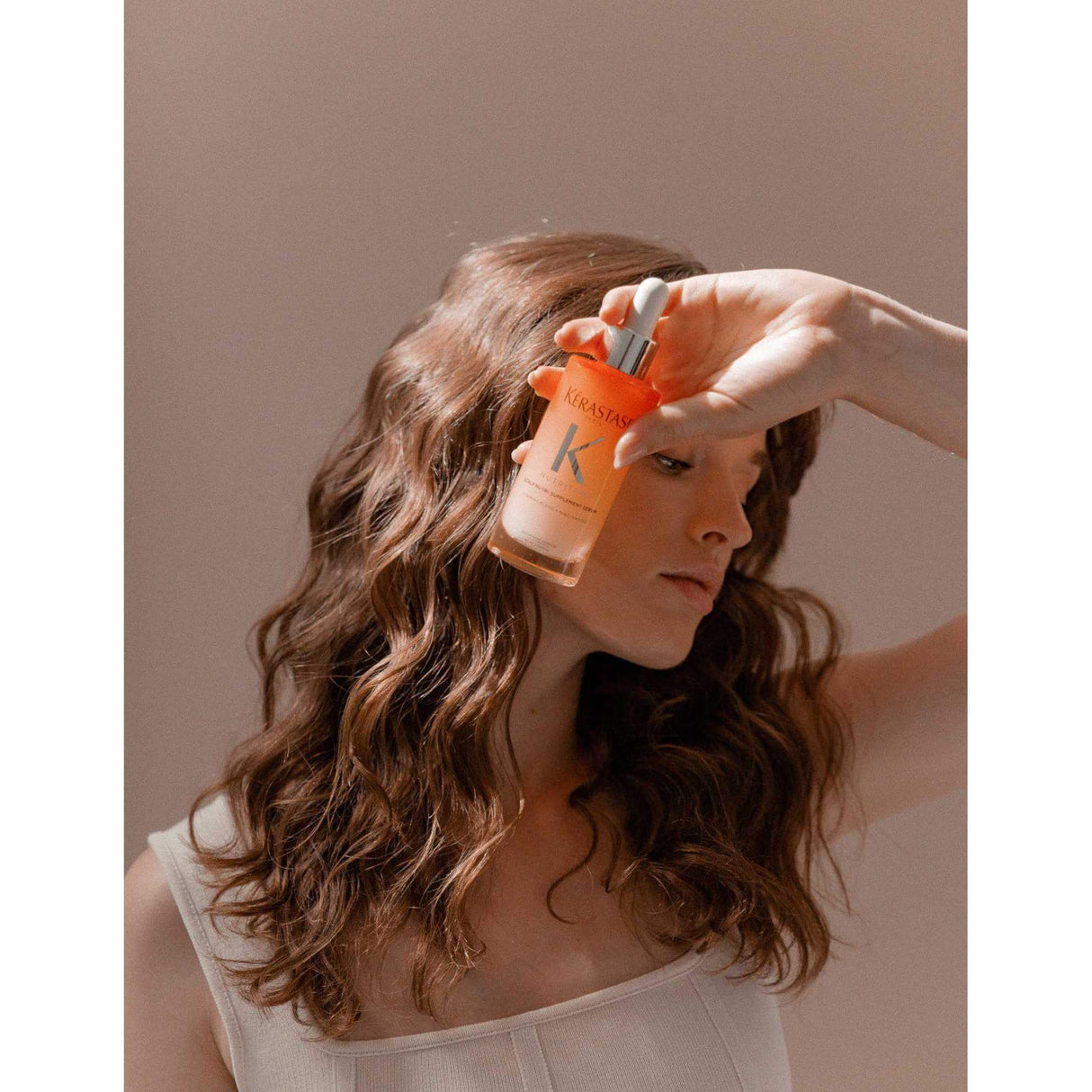 Woman holding Kérastase Nutri-Supplement Scalp Serum bottle with wavy hair, highlighting the product's nourishing benefits.