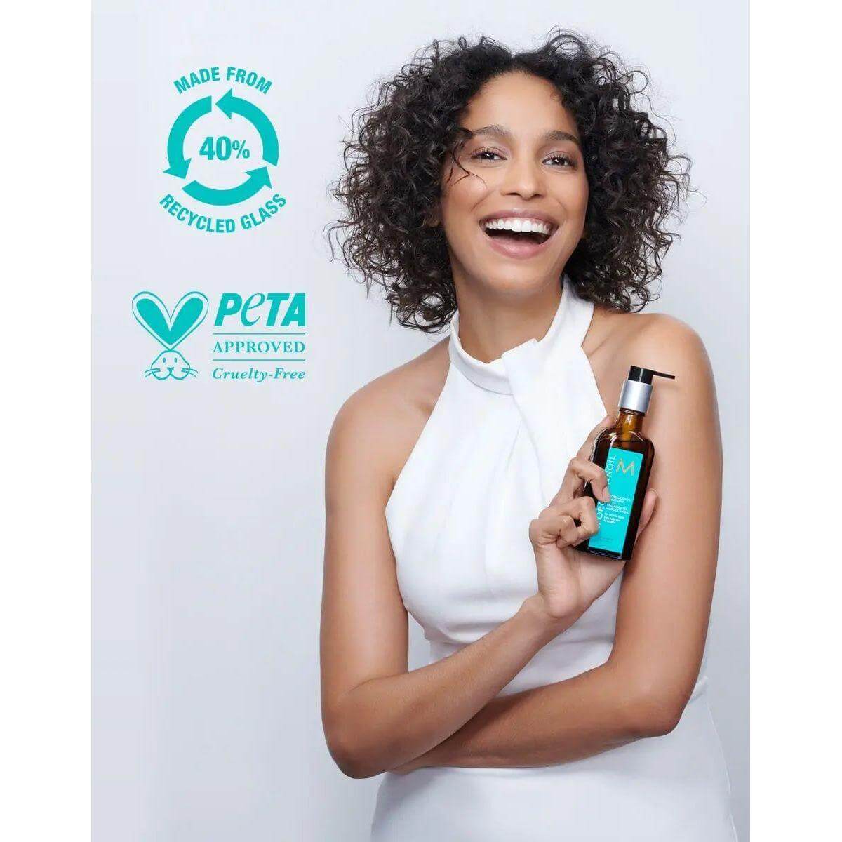 Woman holding Moroccan Oil Original Hair Treatment bottle, featuring PETA approval and 40% recycled glass packaging.