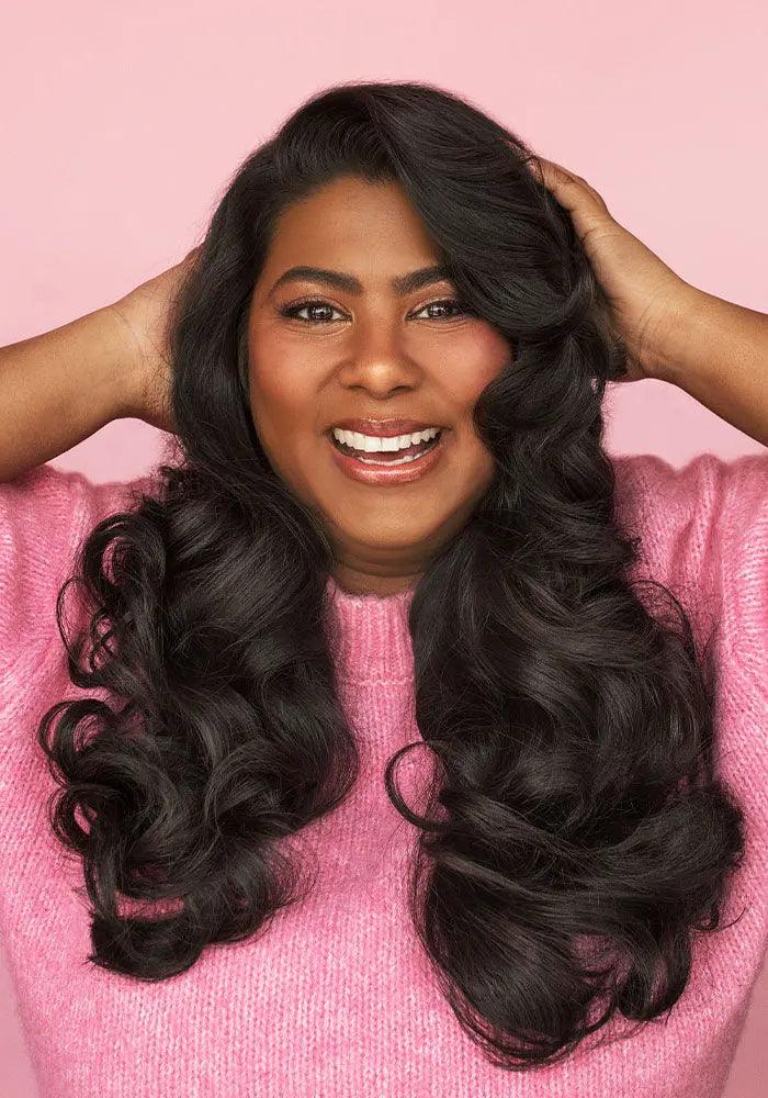 Woman with voluminous curly hair smiling in a pink sweater, showcasing the effect of PUFF.ME Volume Glitz Kit for shiny, soft hair.