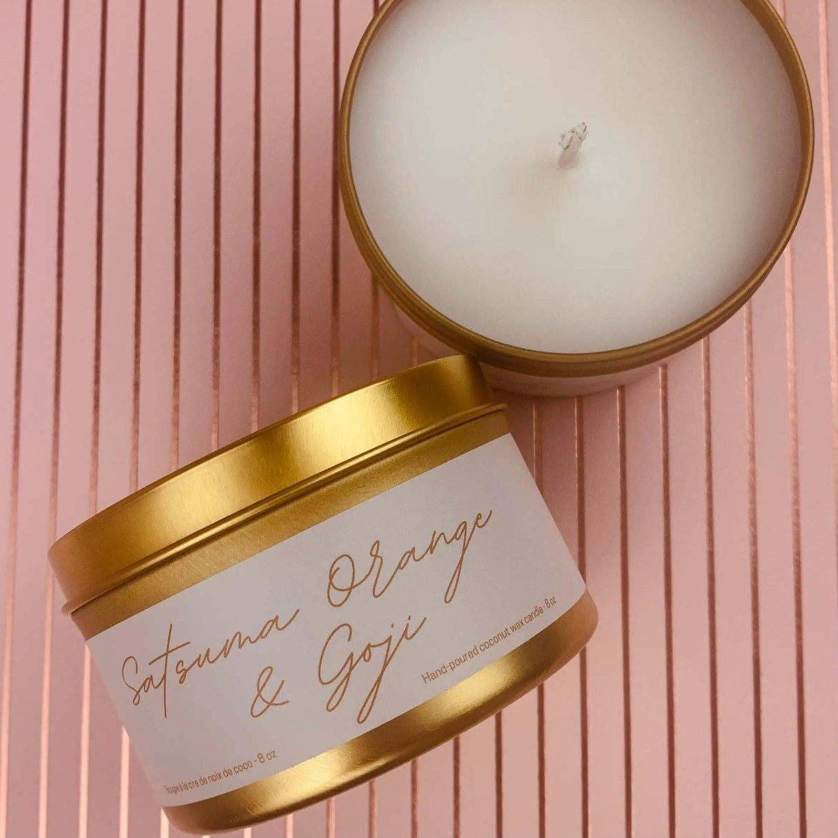 Satsuma Orange & Goji candle in a gold tin placed on a pink striped background.