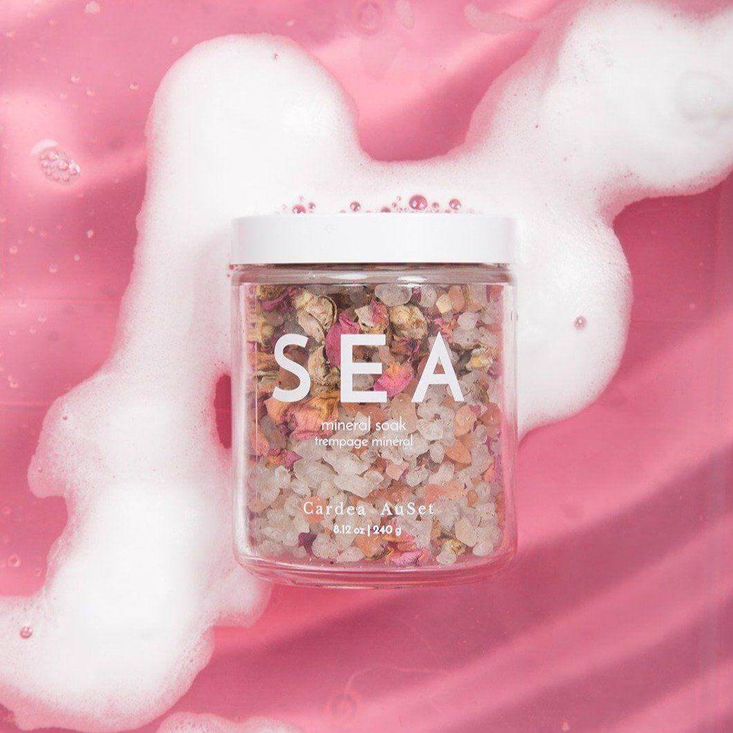 Glass jar of Sea Mineral Soak with Himalayan and Dead Sea salts, rose petals, in a pink bath setting.