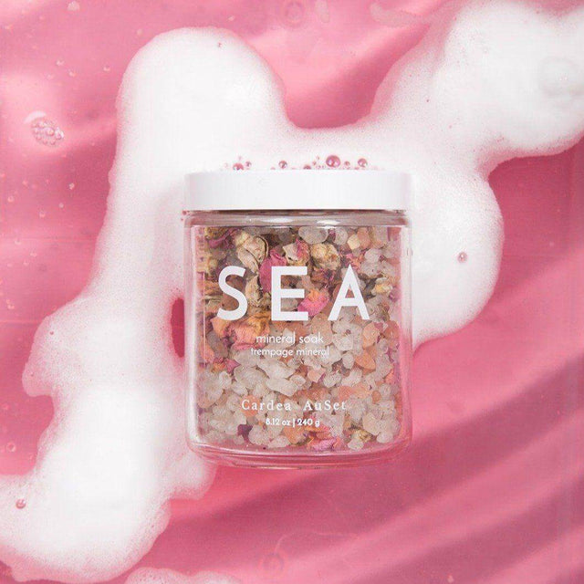 Glass jar of Sea Mineral Soak with Himalayan and Dead Sea salts, rose petals, in a pink bath setting.