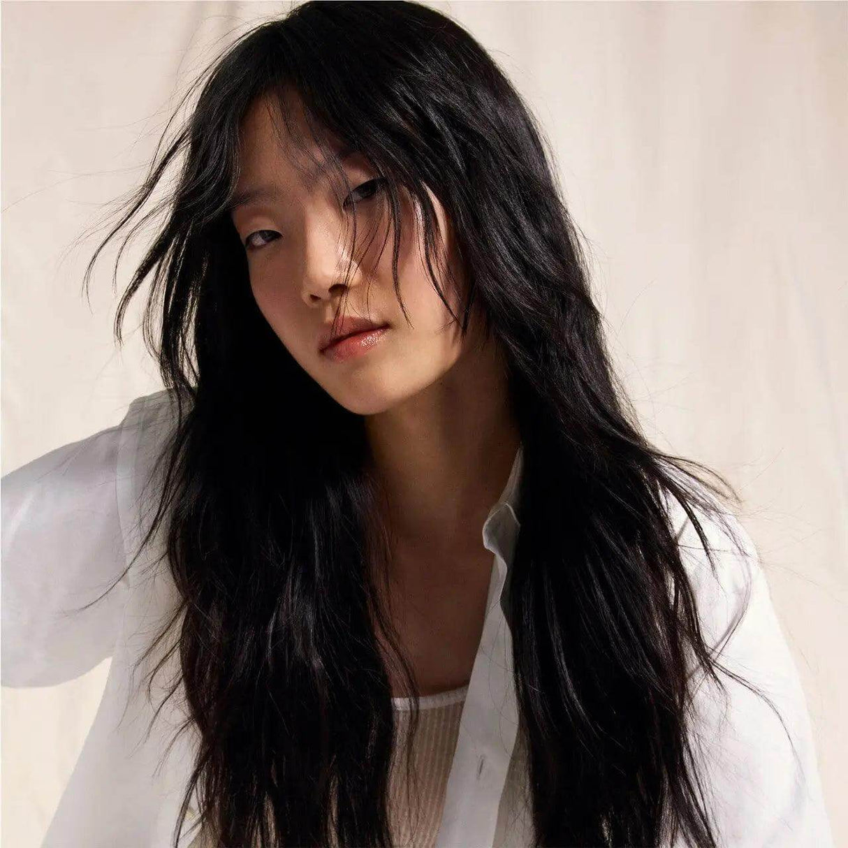 Woman with long, wavy black hair wearing a white open shirt, posing against a neutral background.