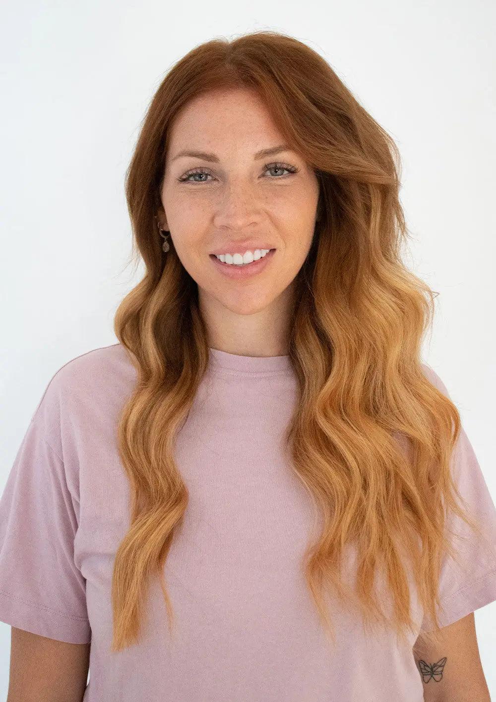 Woman with long wavy hair wearing a light pink t-shirt, smiling against a white background.