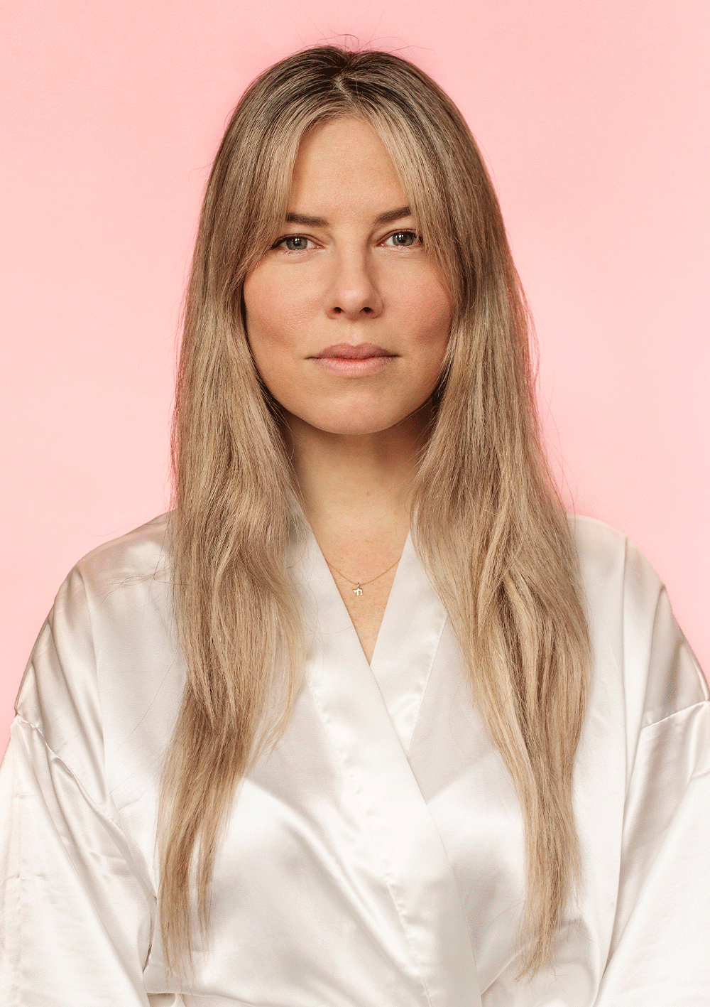 Woman with long blonde hair in a white robe against a pink background.