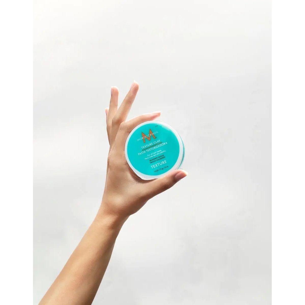 Hand holding Moroccanoil Texture Clay with strong hold and matte finish for versatile hair styling on short and long hair.