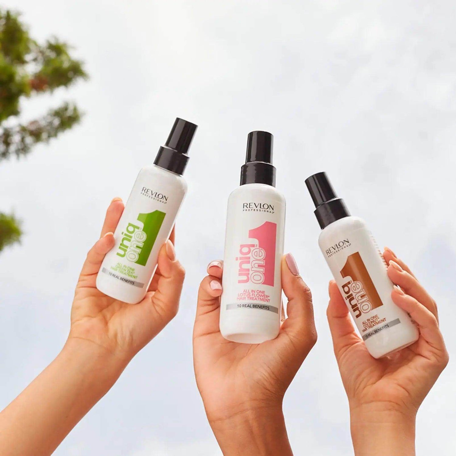 Three bottles of UniqOne Hair Treatment, showcasing different fragrances, held against a clear sky background.