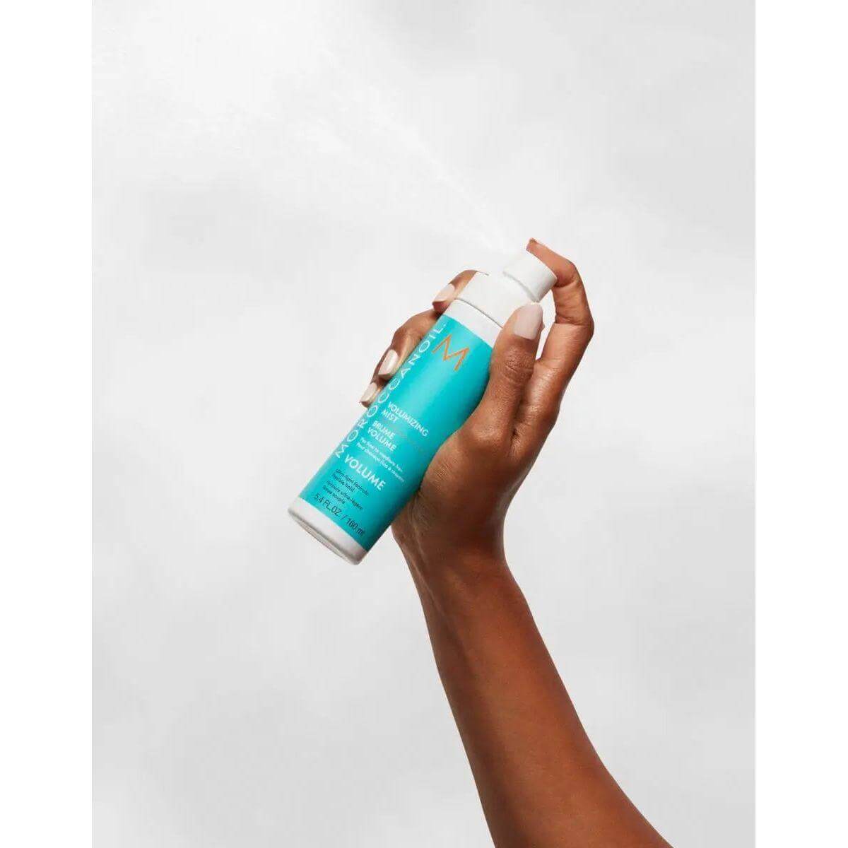 Hand holding Volumizing Mist spray bottle for enhancing hair volume and texture with argan oil and rice amino acids.