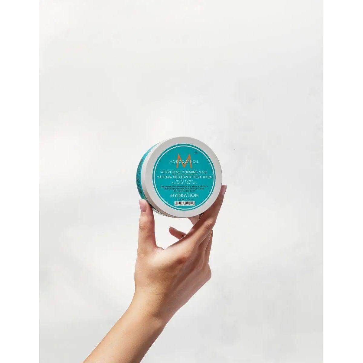 Hand holding Moroccanoil Weightless Hydrating Mask container against white background.