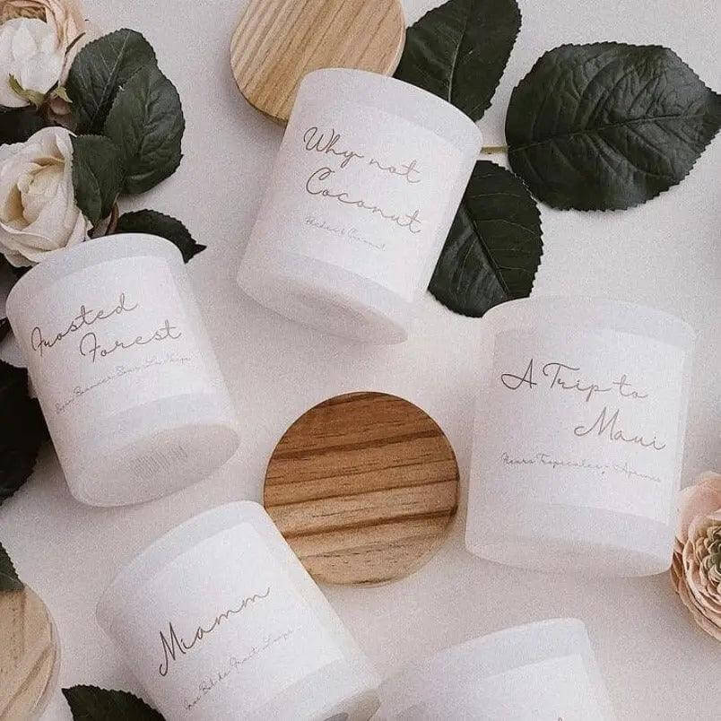 Assorted scented candles with labels, including Why Not Coconut, surrounded by leaves and wooden lids.