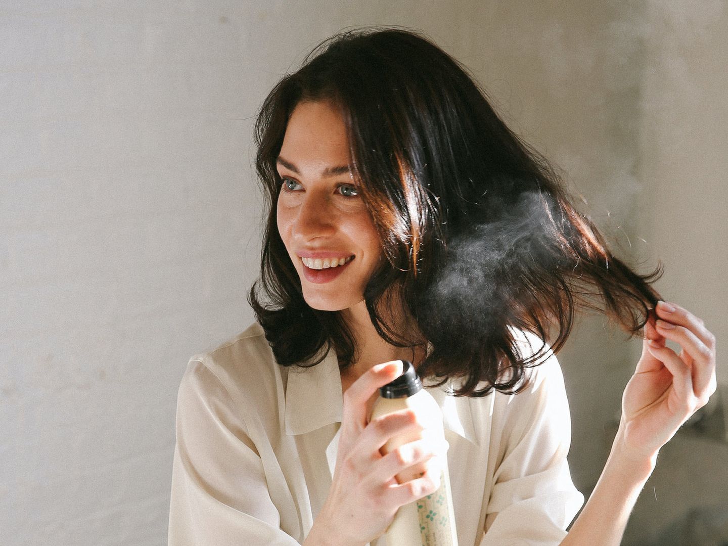 Woman spraying hair product, smiling, indoors, natural light.