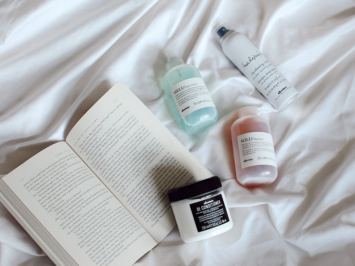 Skincare products and an open book on a white bedspread.