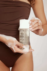 Woman in brown swimsuit using a luxurious body wash, part of the Body Essentials Trio, perfect for sunless tanning preparation and care.