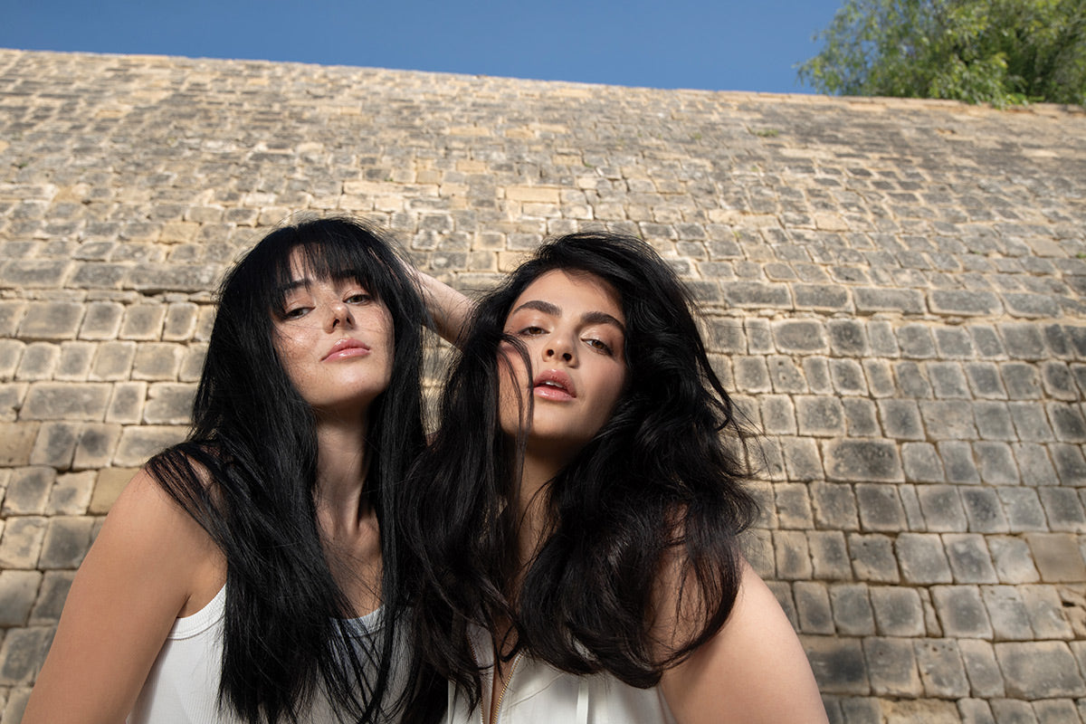 Two women with long dark hair styled with Neu Styling Flexible Hairspray, posing outdoors against a stone wall.