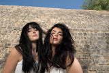 Two women with long dark hair styled with Neu Styling Flexible Hairspray, posing outdoors against a stone wall.