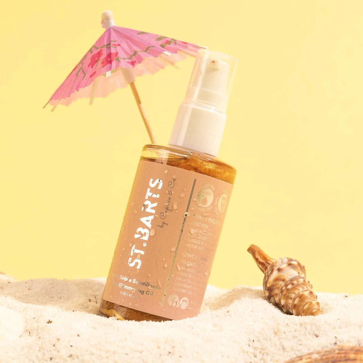 St. Barts Dry Shimmering Oil with tropical scents, on beach sand with seashell and mini umbrella, enhances skin with bronze glow.