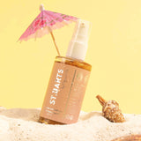 St. Barts Dry Shimmering Oil with tropical scents, on beach sand with seashell and mini umbrella, enhances skin with bronze glow.